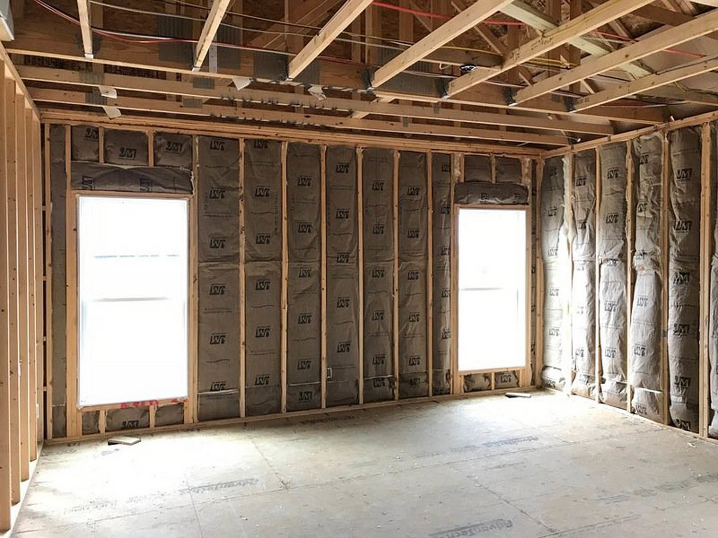 Room under construction with exposed wooden framing, wall filled with insulation, concrete floor, and window letting in natural light