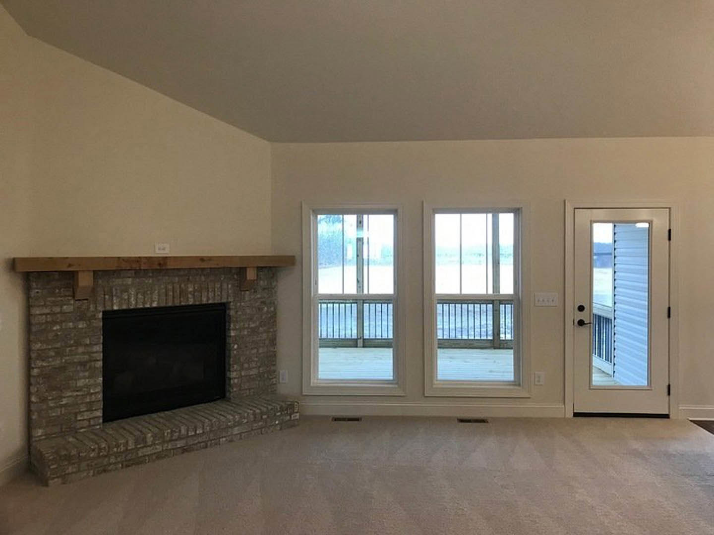 Carpeted living room featuring a black fireplace set in brick with a wood mantel, white door with window, two windows with railing, and wall vent.
