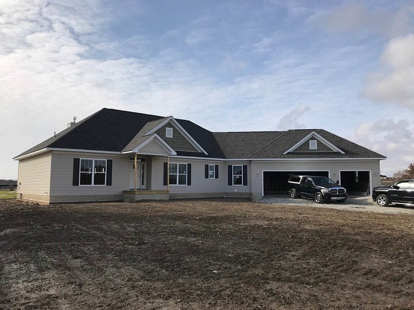 Black pickup truck parked in gravel driveway in front of modern white house with gabled roof, covered porch, large windows, and grassy yard under cloudy sky.