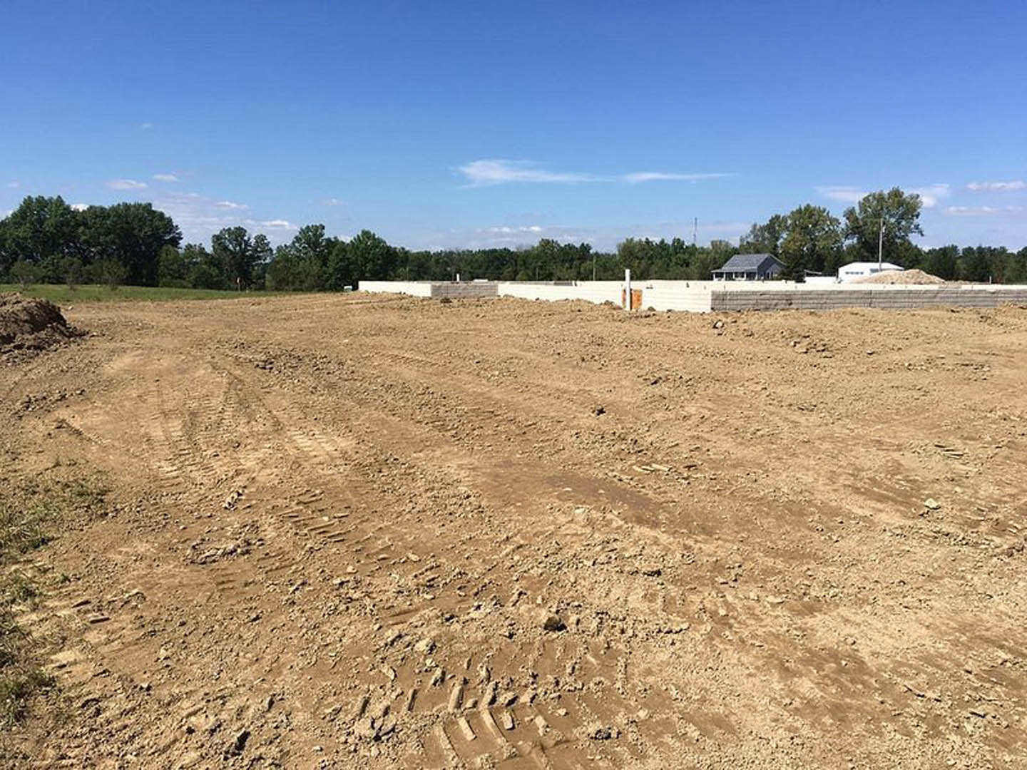 Expansive dirt field with tire tracks, scattered piles of soil, green-leaved trees lining the background, blue sky with clouds overhead