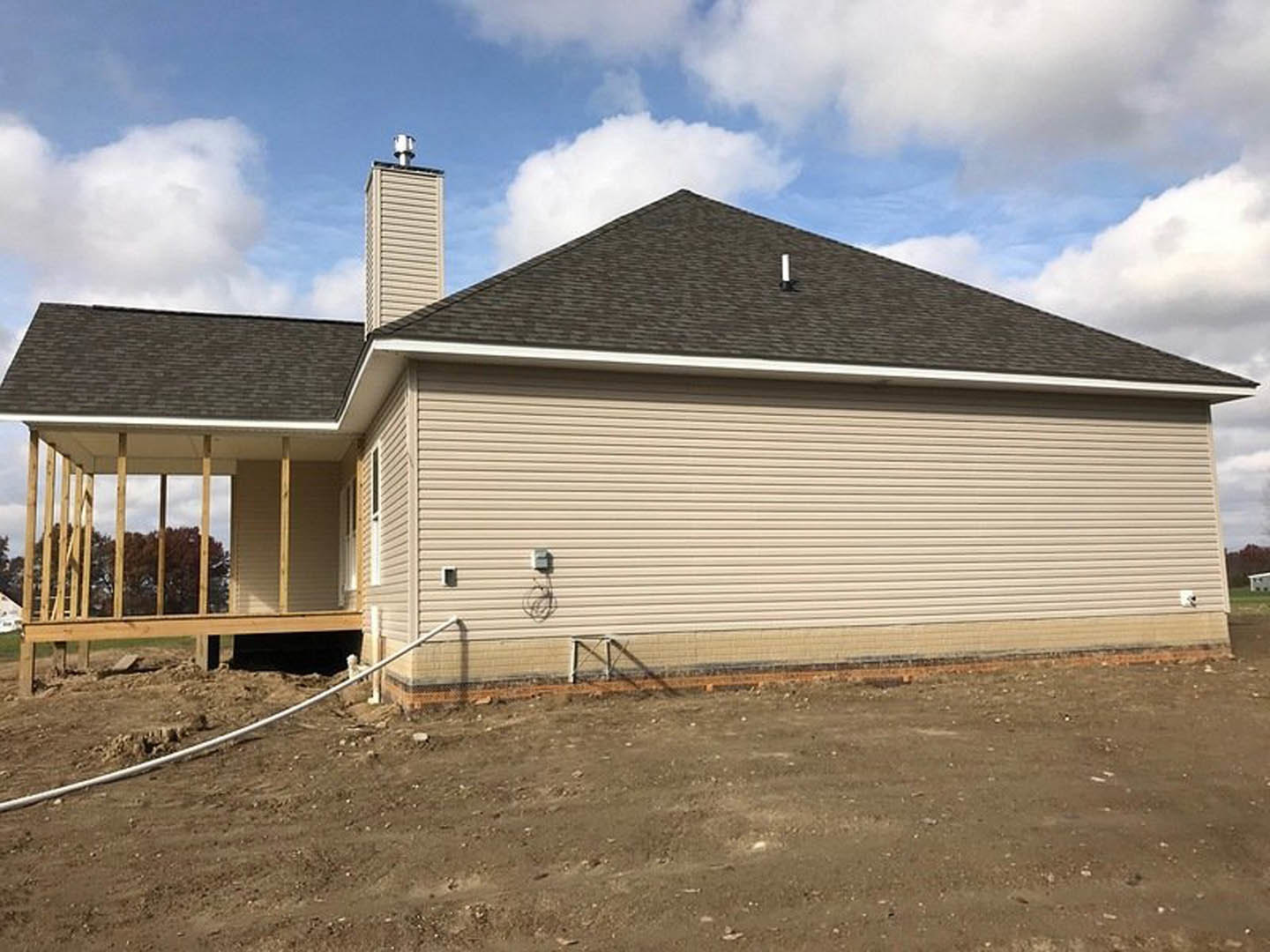 Two-story house with gray siding, covered front porch, white railing, wooden fence, brick chimney, dirt yard, and scattered construction materials under partly cloudy sky