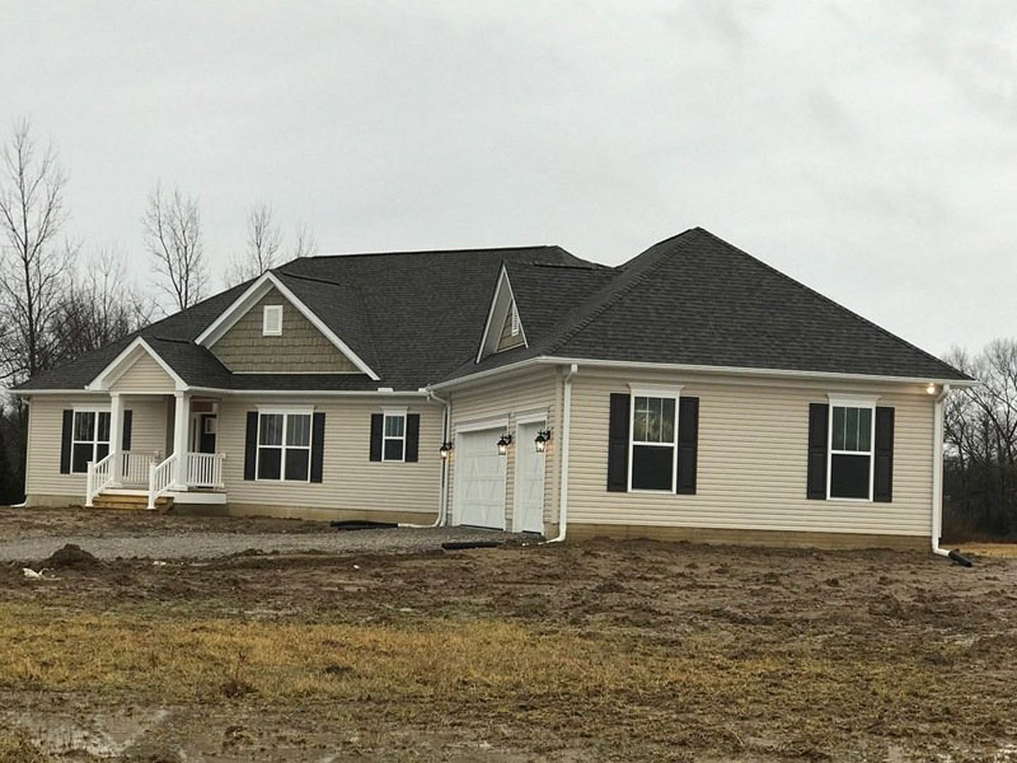 Two-story house with gray siding, large paved driveway, attached garage with white door and exterior lights, white porch railing, white-framed windows, and landscaped yard with