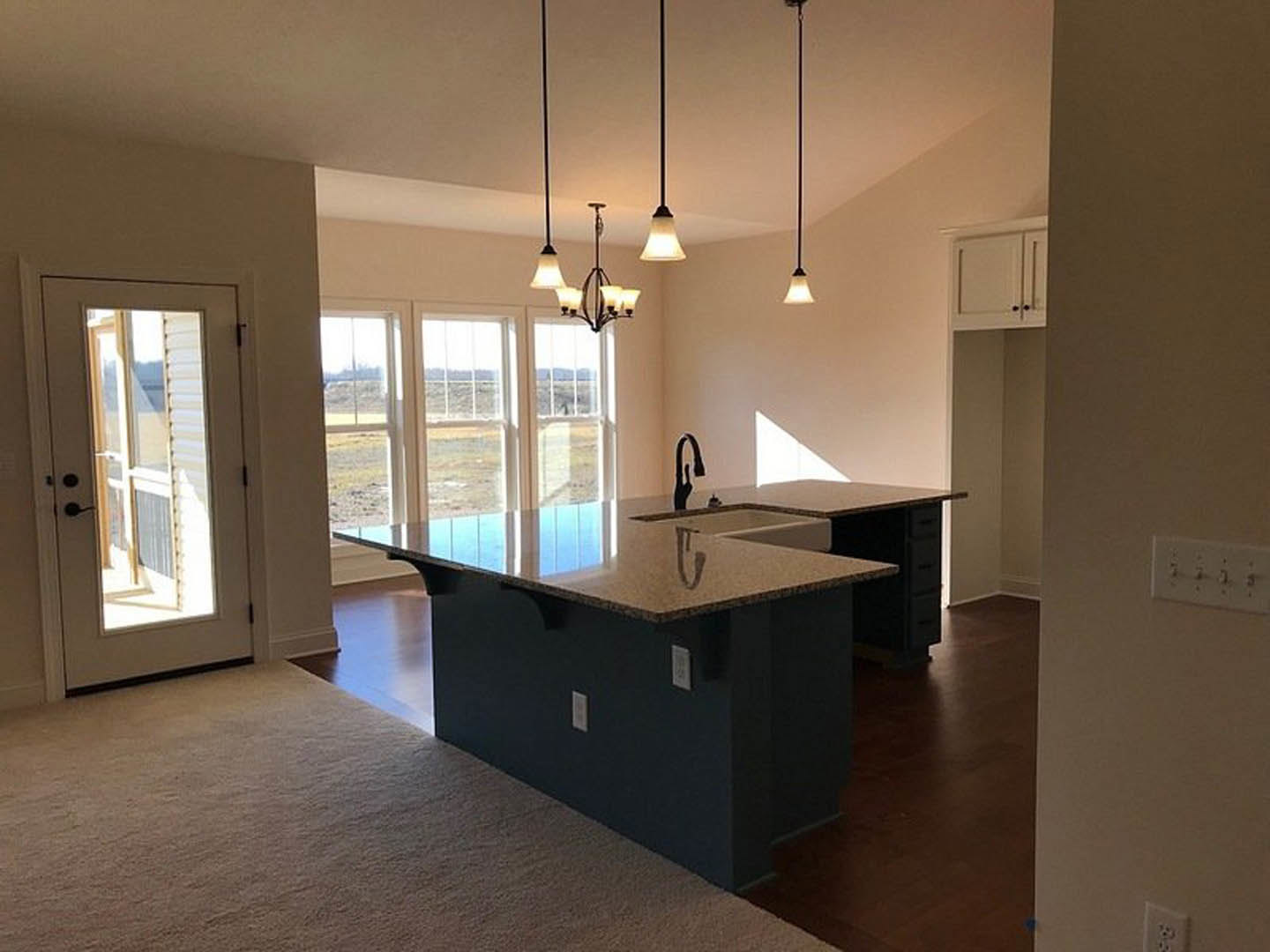 Bright kitchen featuring a central island with built-in sink, white cabinetry, stone countertops, large window overlooking green lawn, wood flooring, ceiling chandelier, and open