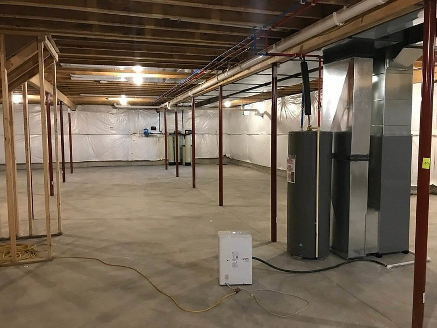 Exposed steel and black pipes run along ceiling and walls in unfinished room with wooden framing, white electrical box, and white plastic wall covering.