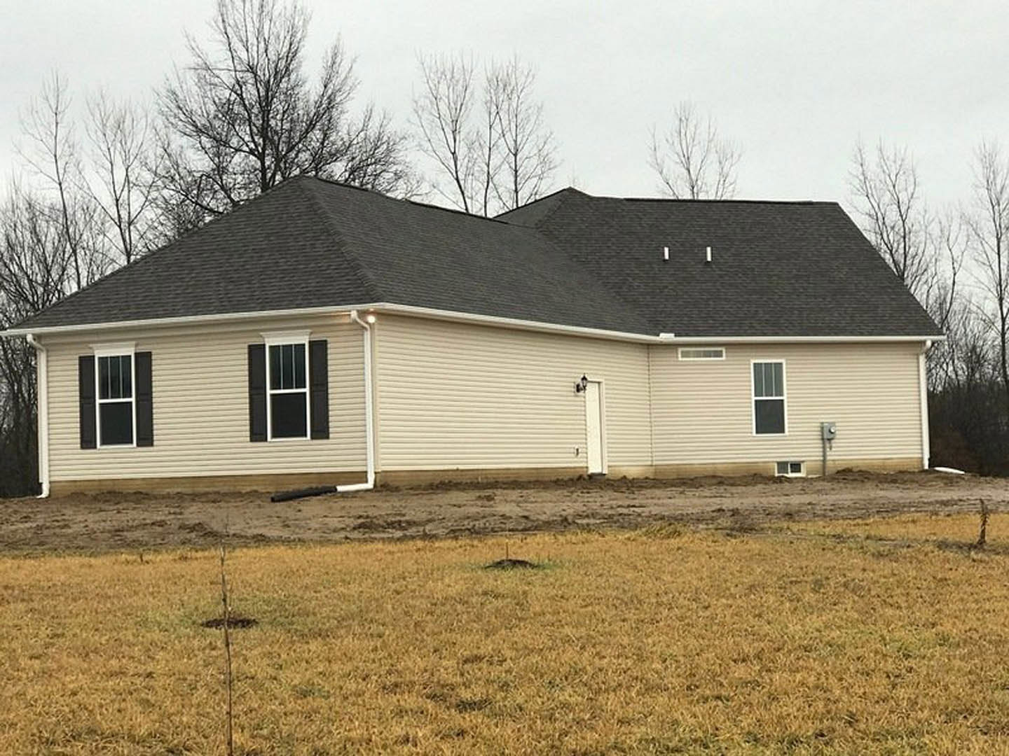 Two-story farmhouse with white roof and white-framed windows, light siding, dirt patch and grassy area in front, small tree and stick in yard under blue sky