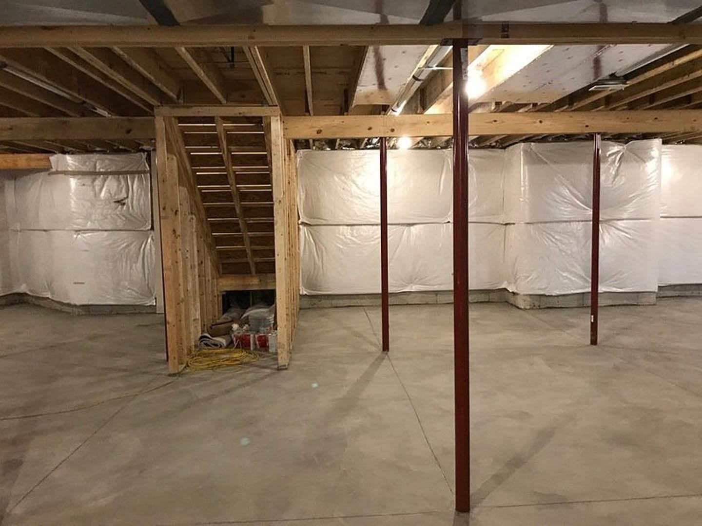 Exposed wooden ceiling beams above a white plaster wall, concrete floor, and stacked white plastic bags; wood shelving along one side.