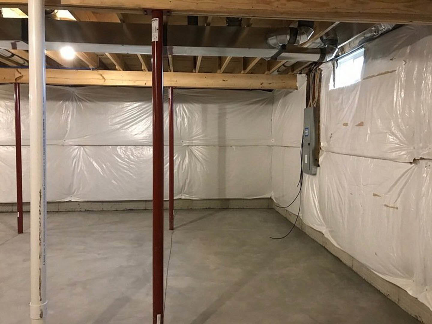White plaster wall with exposed metal support pole, concrete floor with visible wiring, large window allowing bright natural light, ceiling partially wrapped in white plastic