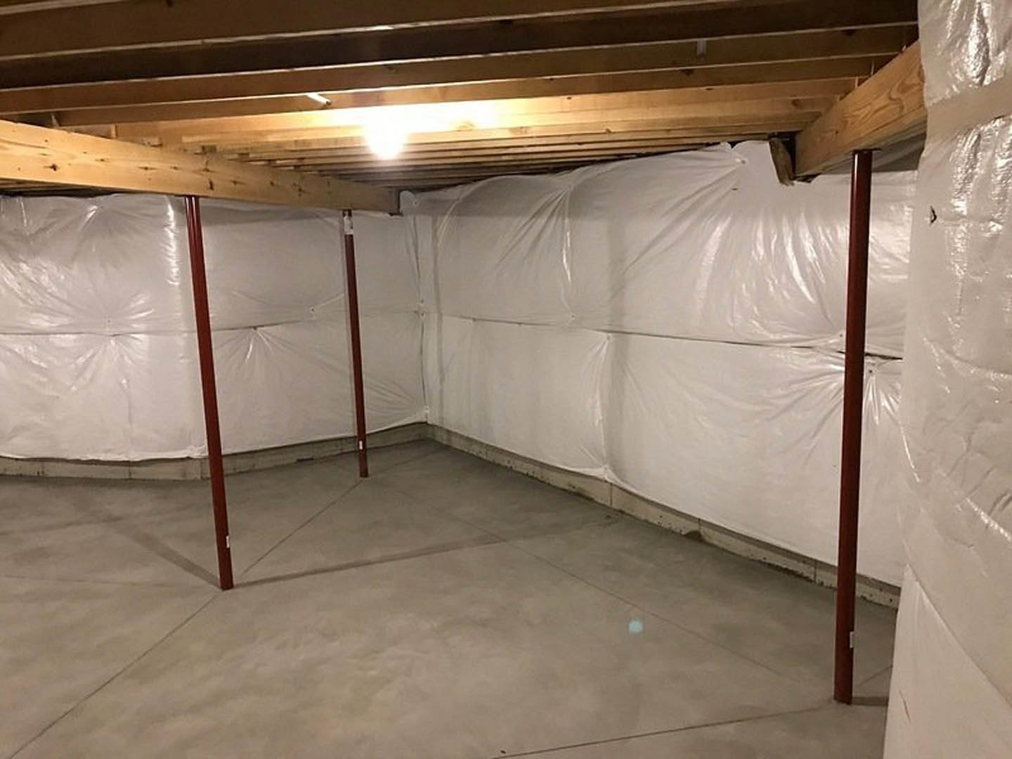 Basement with concrete floor, exposed support pole, and walls covered in white plastic sheeting; overhead light illuminates ceiling and insulation.
