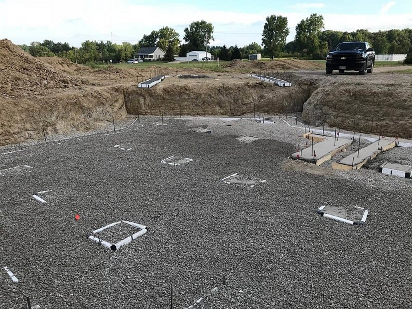 Black-roofed house beside gravel pile with white square objects, black truck parked on dirt, green-leafed tree in foreground