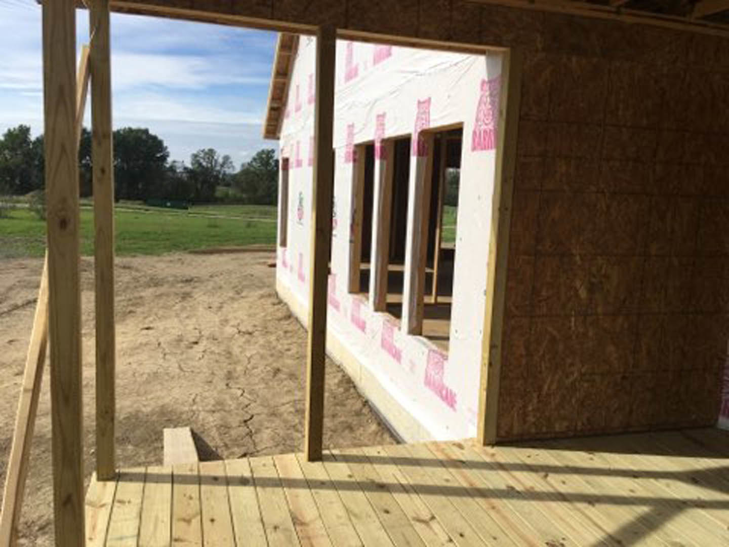 Wood-framed house under construction with exposed wooden deck, pink tape marking boundaries, grassy field and tree in background, partial view of fence in foreground