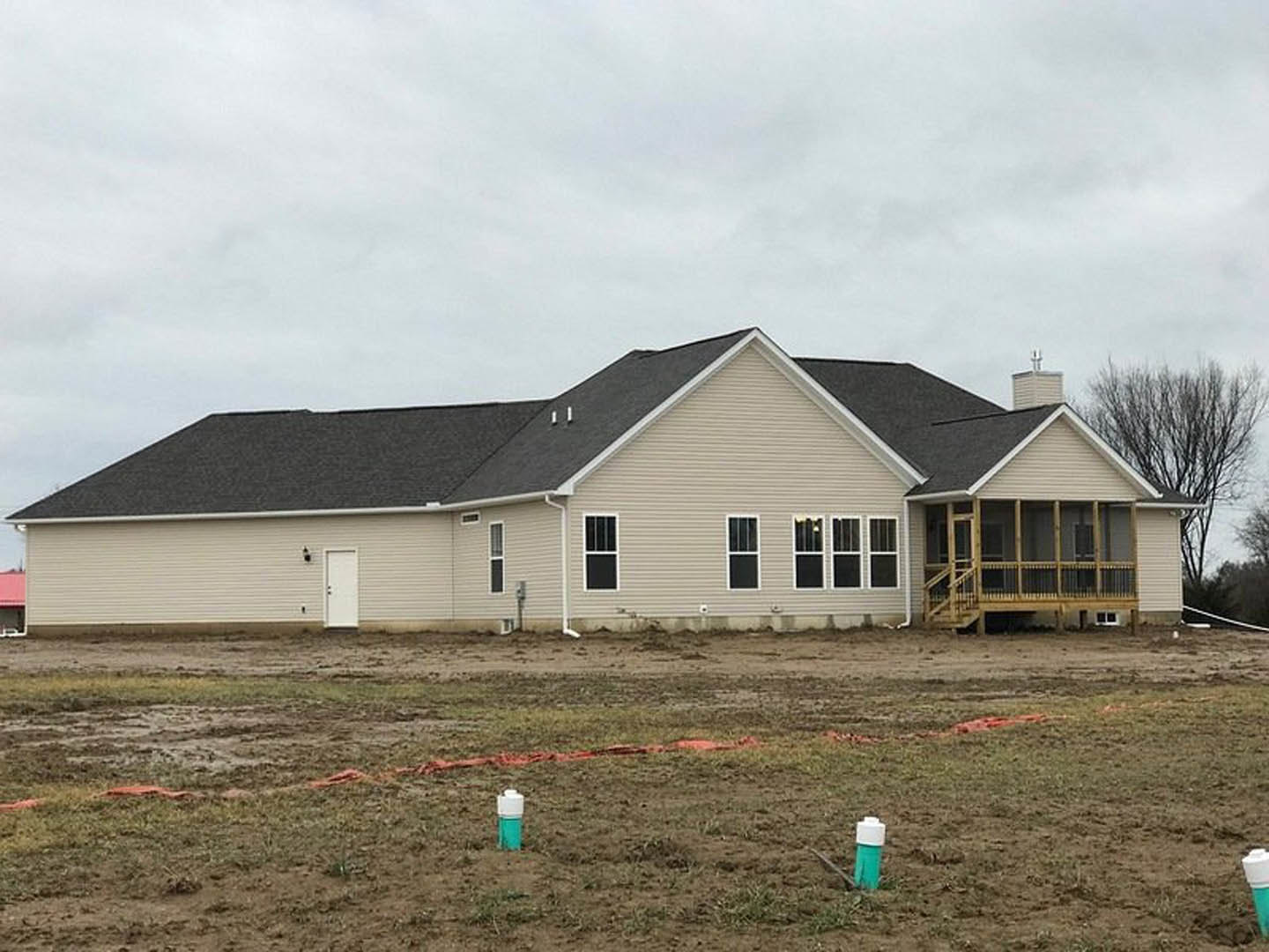 Two-story farmhouse with black roof, white siding, covered front porch, white door with black handle, grassy yard, small tree, and green and white container near dirt walkway