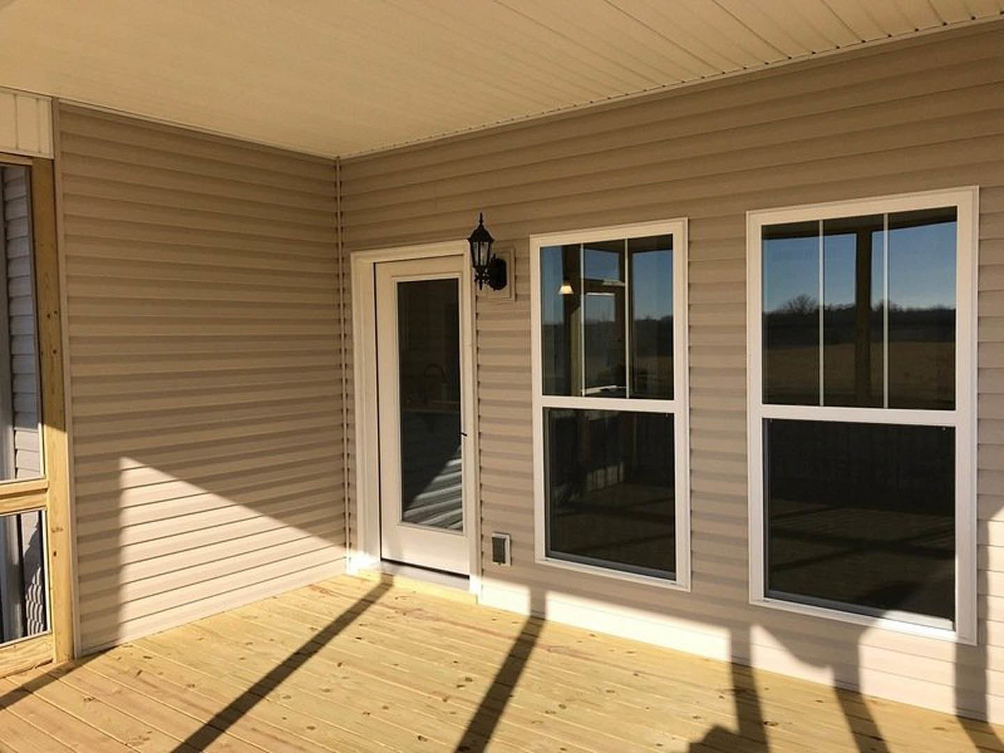 White siding house with covered front porch, two large windows, black wall-mounted light fixture, wooden floorboards, and sunlight reflecting off glass.