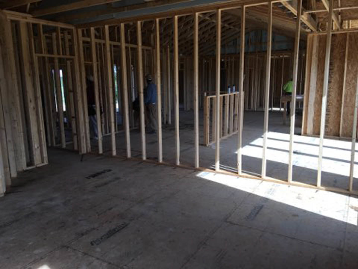Framed interior under construction with exposed wooden beams, concrete floor, and workers installing materials
