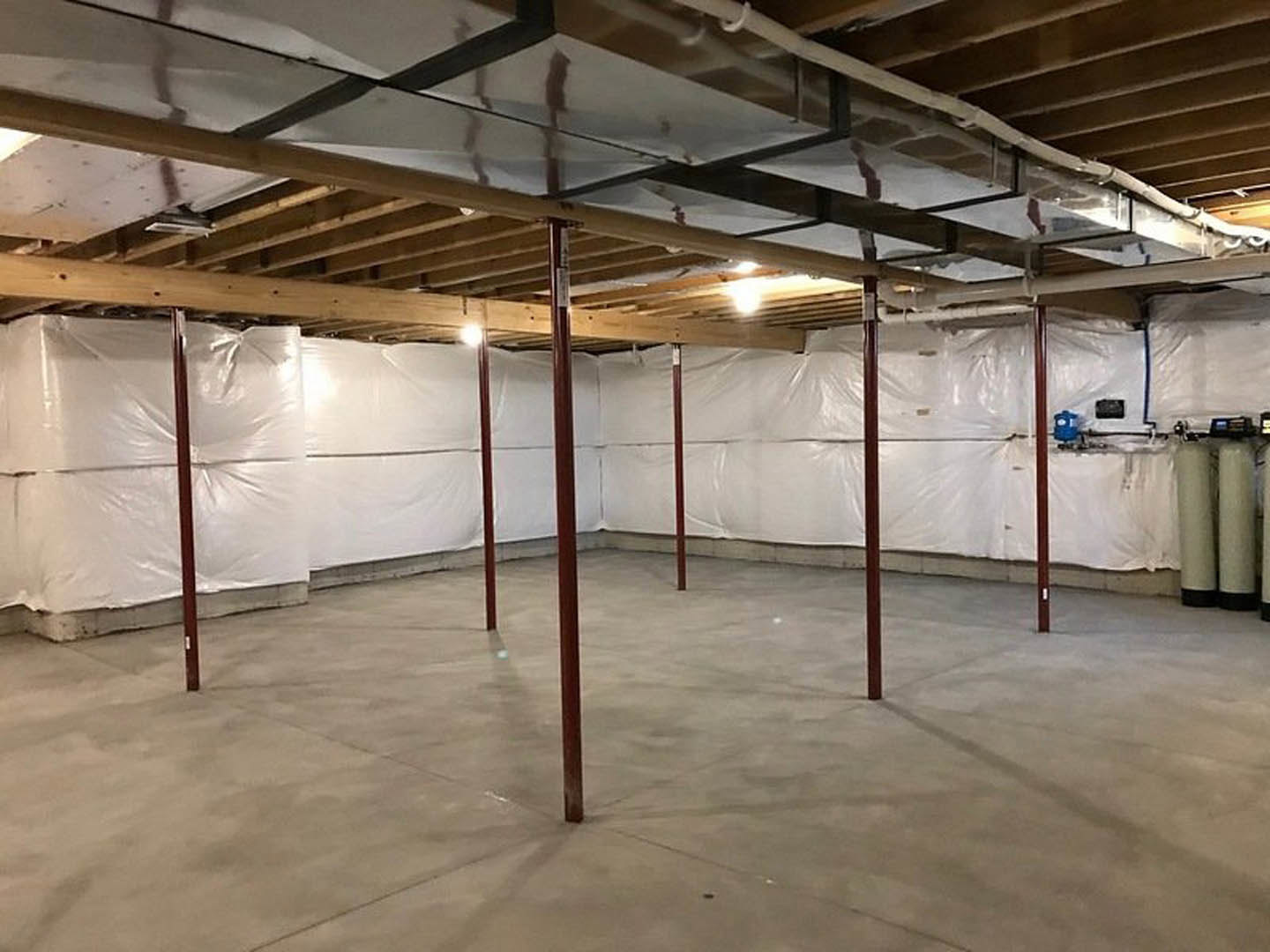 Unfinished room with white plastic coverings on walls, exposed concrete floor with red poles, ceiling featuring pipes and a metal beam, bright light reflecting on surfaces