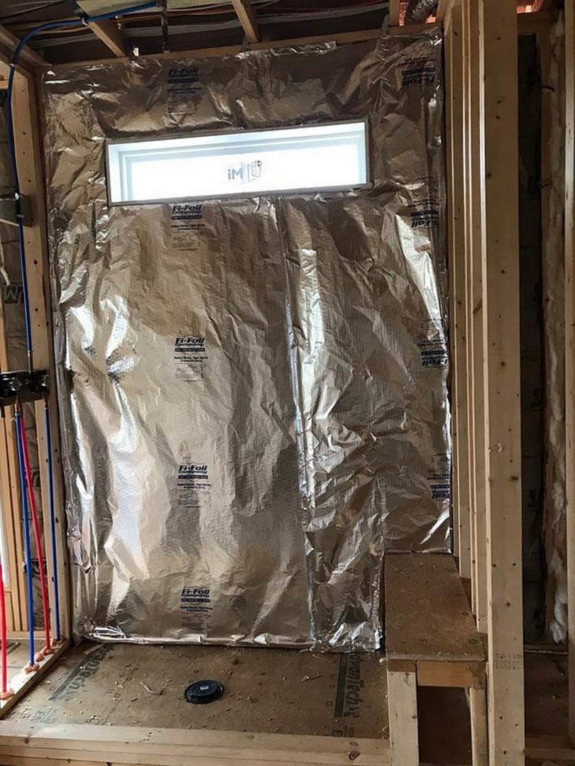 White interior door with foil covering, small window, and wall-mounted light fixture; nearby shelf holds a stack of books, black drain visible on brown flooring.