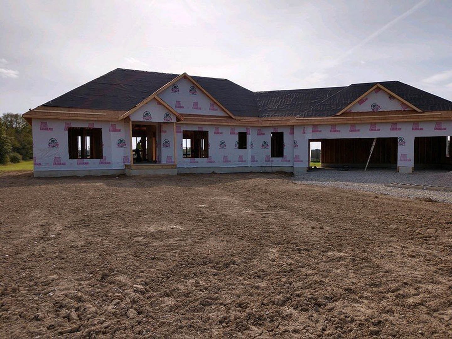 Framed custom home under construction with exposed wood, pink tape marking entry, dirt lot in foreground, windows installed, cloudy sky overhead