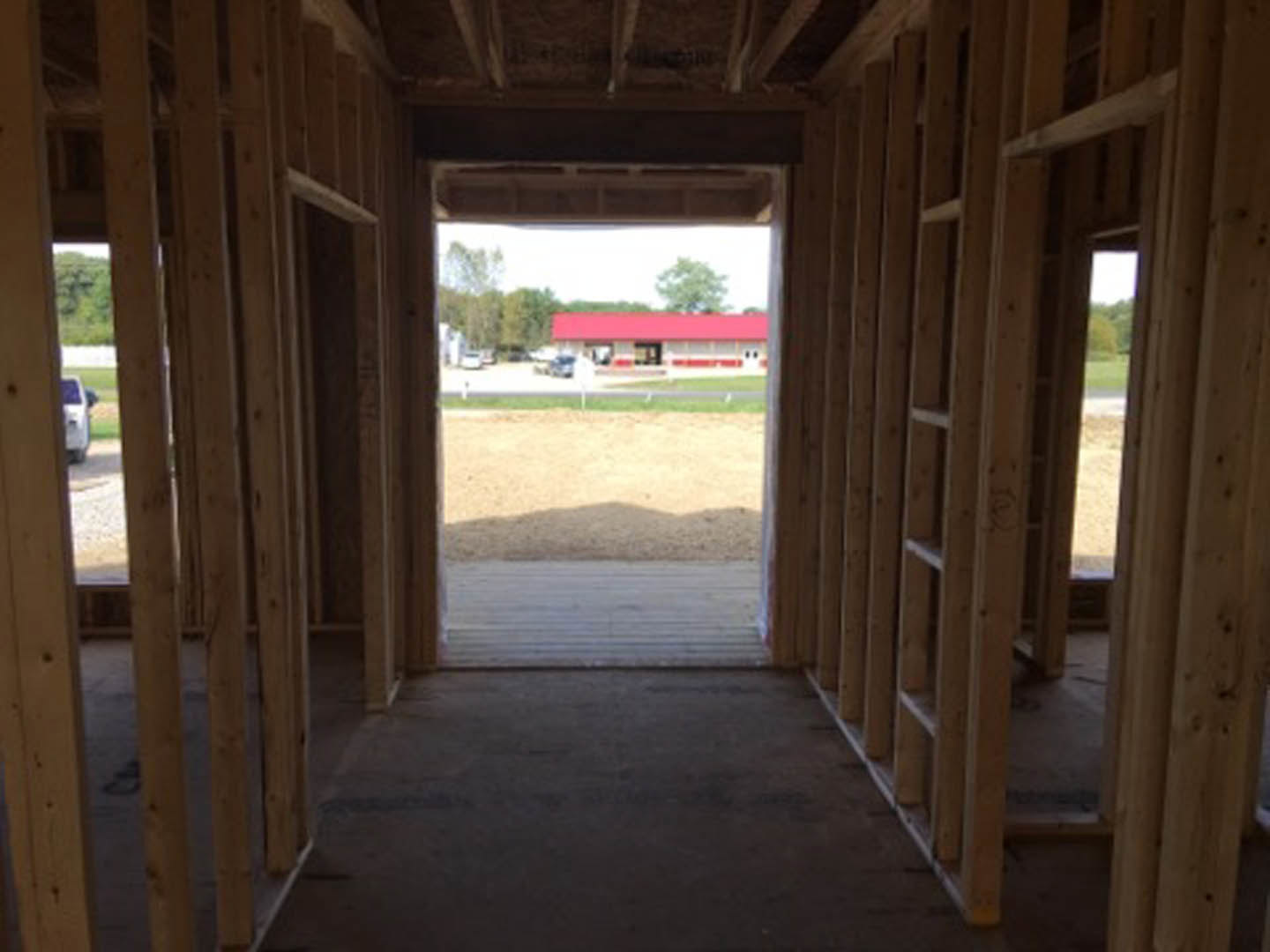 Framed house under construction with exposed lumber, open doorway, and red building visible in the background