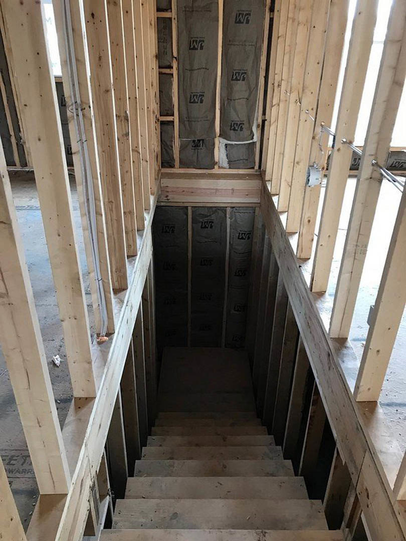 Wooden staircase with unfinished handrail and exposed beams in a house under construction, grey composite material on shelving, dark flooring, and visible construction materials.