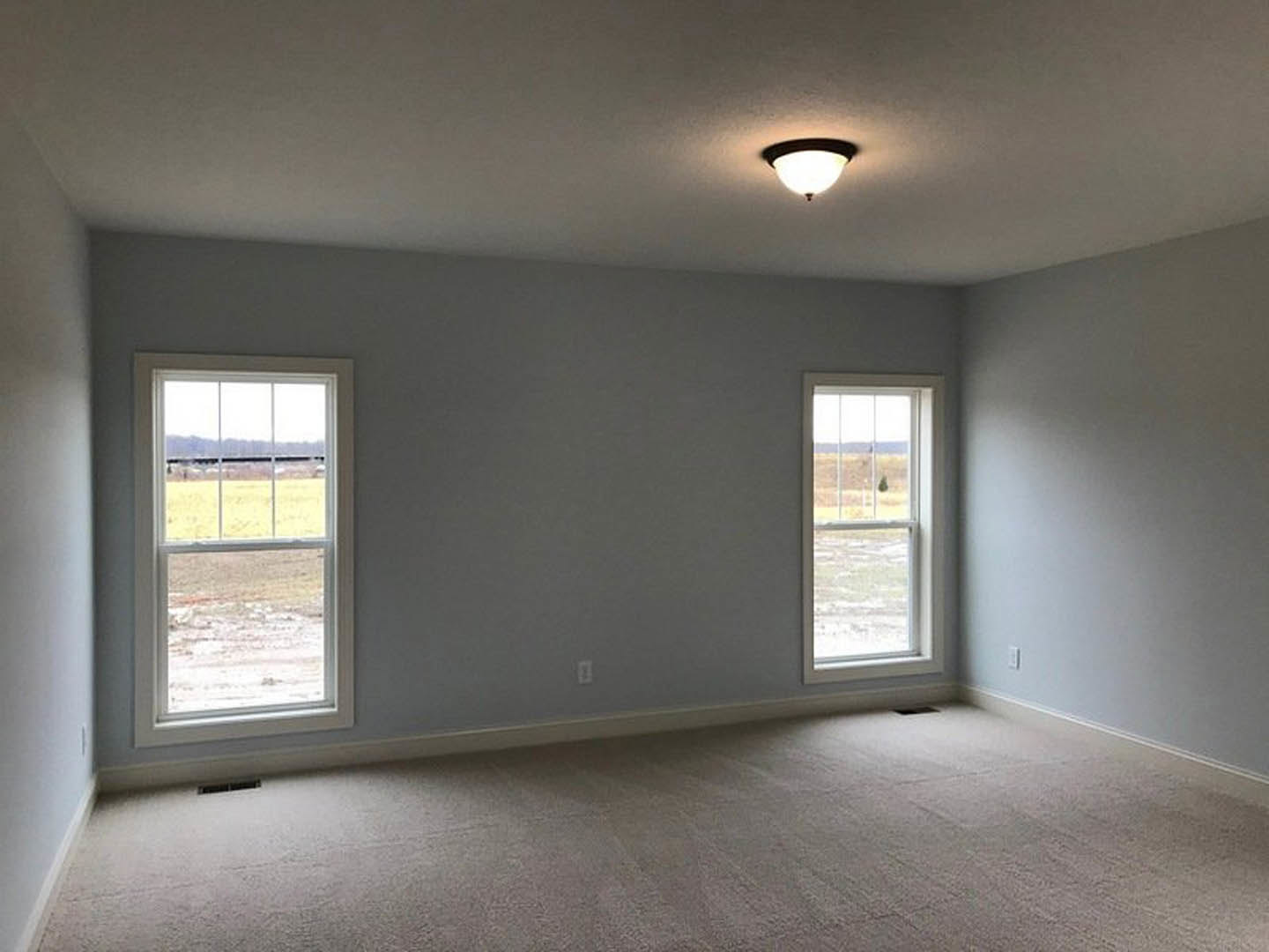 Carpeted room with two large windows, plaster walls, ceiling light fixture, and view of grassy field outside