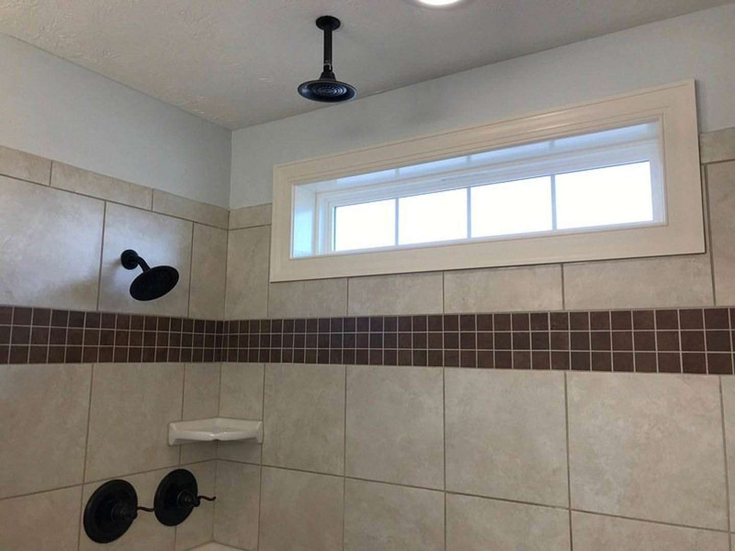Tile shower with a white-framed window, black shower head, black metal plates on the wall, white corner shelf, and ceiling light.