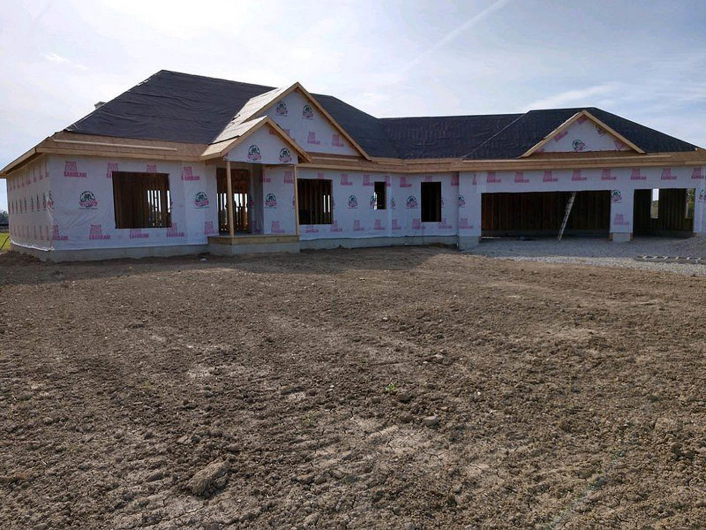 Two-story house under construction with exposed framing, pink tape along the side, surrounded by dirt and construction debris, cloudy sky overhead