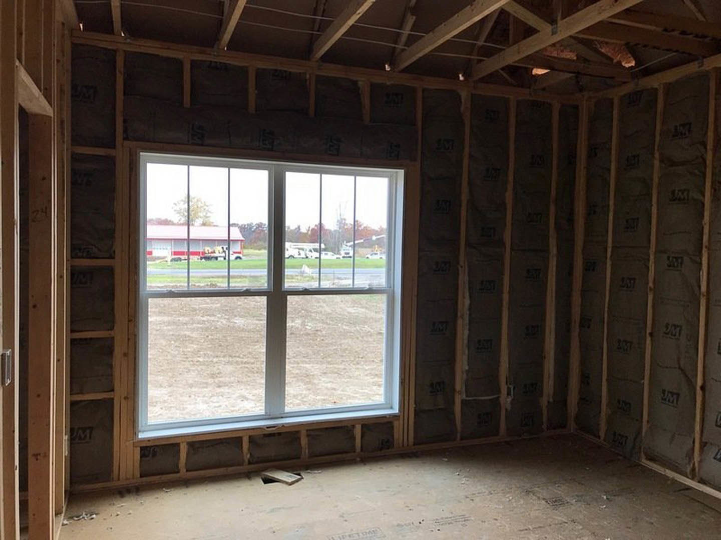 Bright interior room with wood beam ceiling, large window overlooking a field and red-roofed building, concrete floor, and white walls.