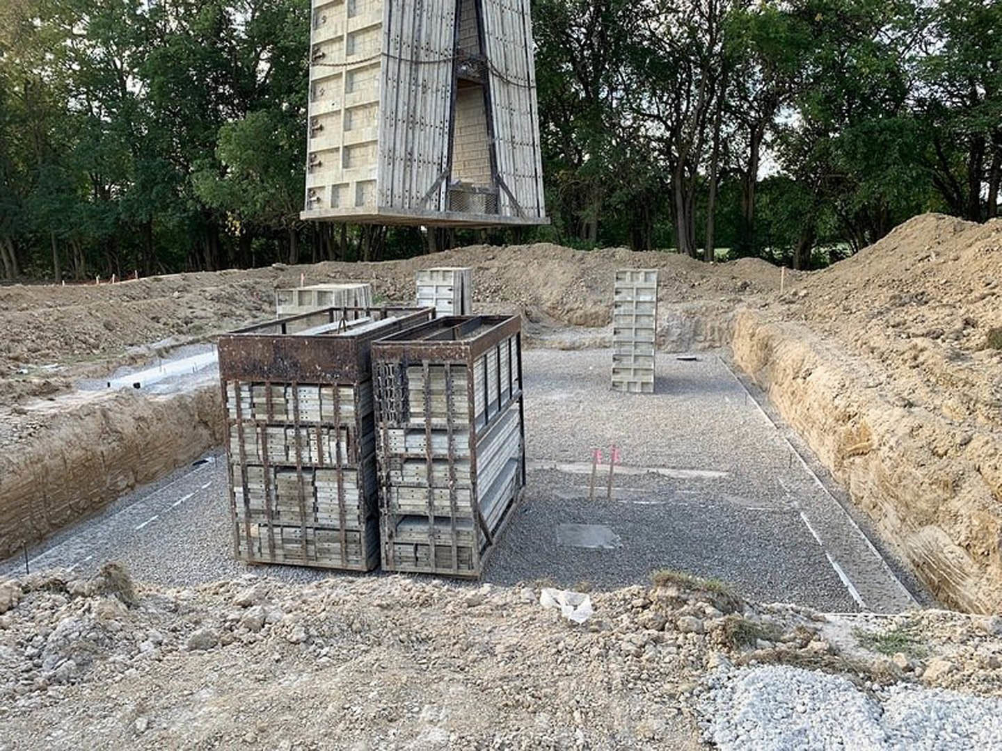 Stone exterior custom home under construction, surrounded by pallets, metal crates, and stacks of shelves; wooden structure with wall opening, trees and plants visible around the