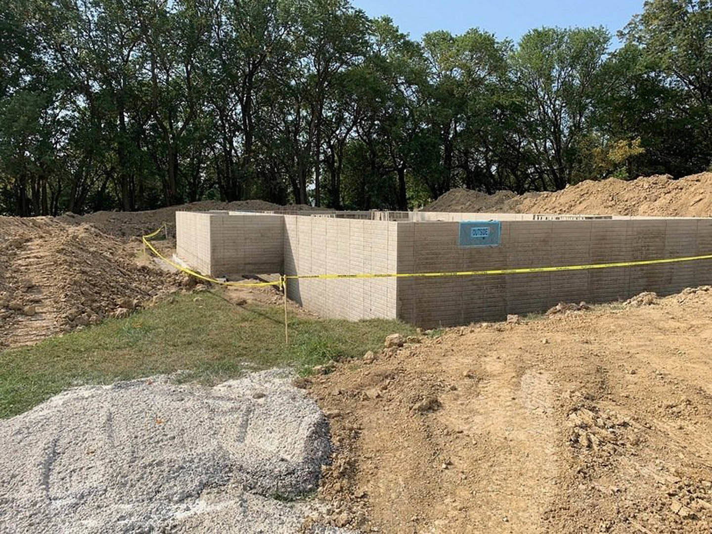 Wood-framed custom home under construction surrounded by dirt, scattered rocks, and mature trees, with yellow caution tape marking the site boundaries.
