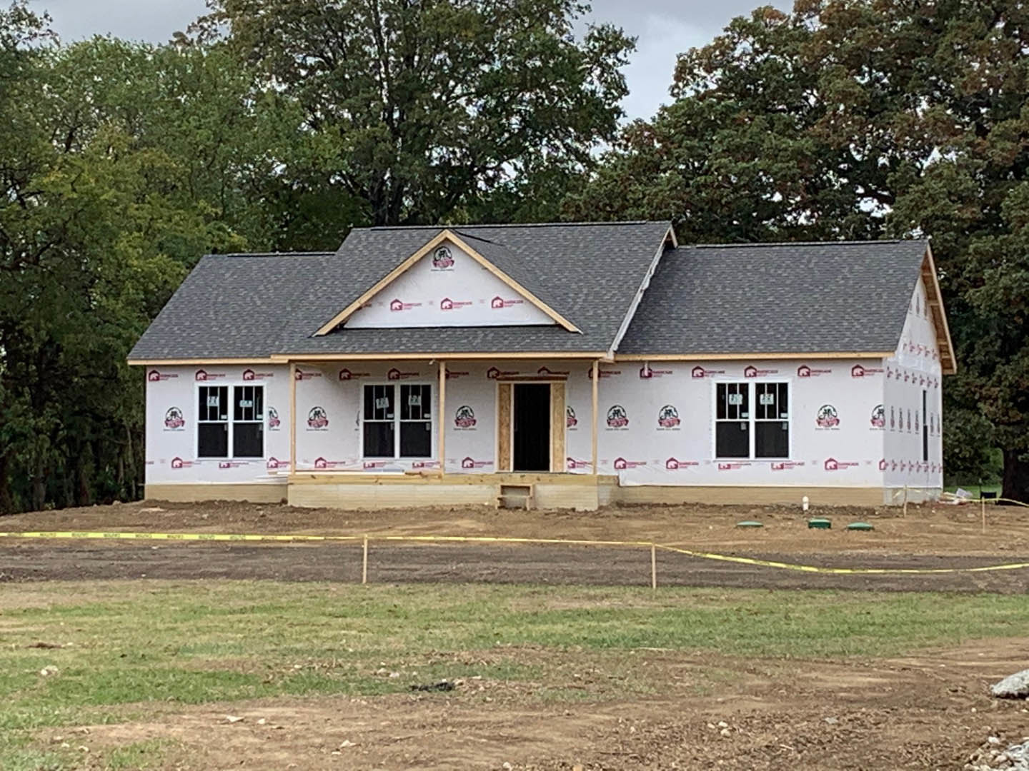 Wood-framed house under construction with exposed roof trusses, surrounded by mature trees and dirt lot
