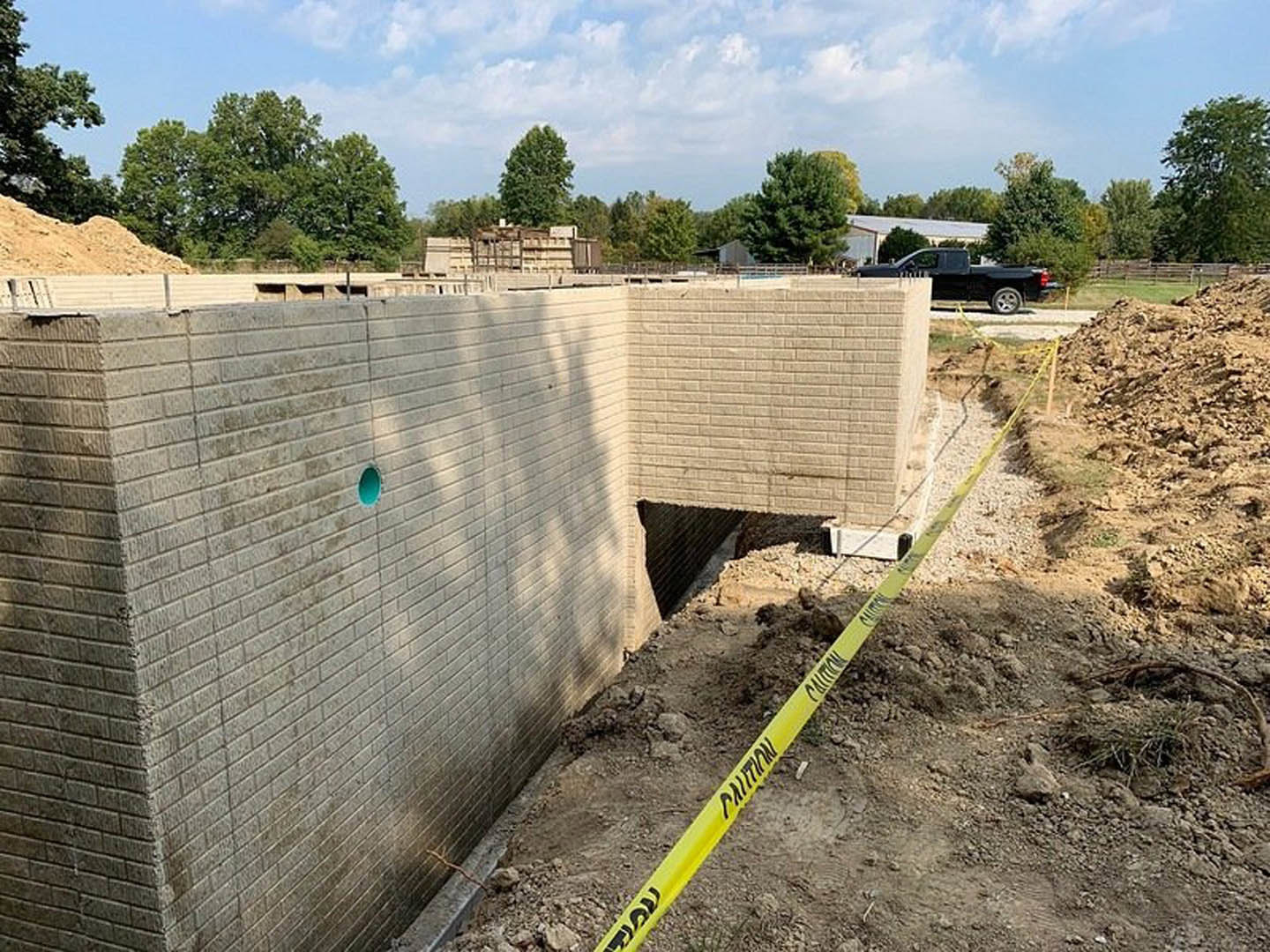 Brick wall under construction with exposed hole, yellow caution tape on ground, black truck parked nearby, blue sky with scattered clouds and surrounding trees