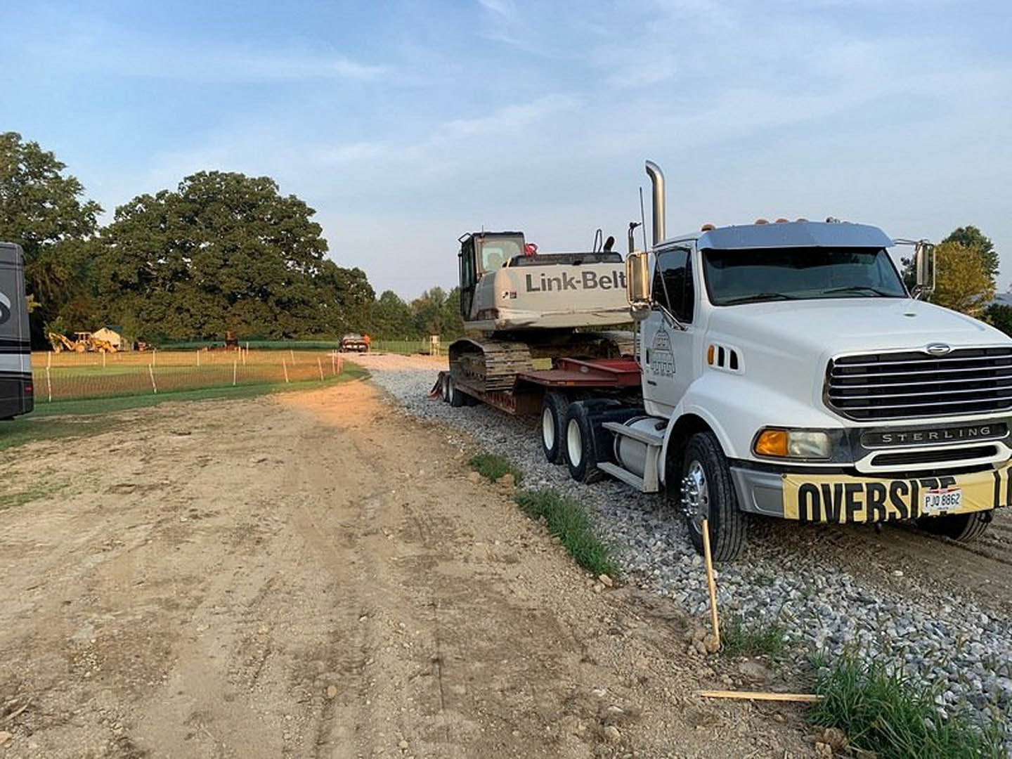 White pickup truck with trailer parked on dirt road beside fenced grassy field, large leafy tree in background, close-up of tire visible in foreground