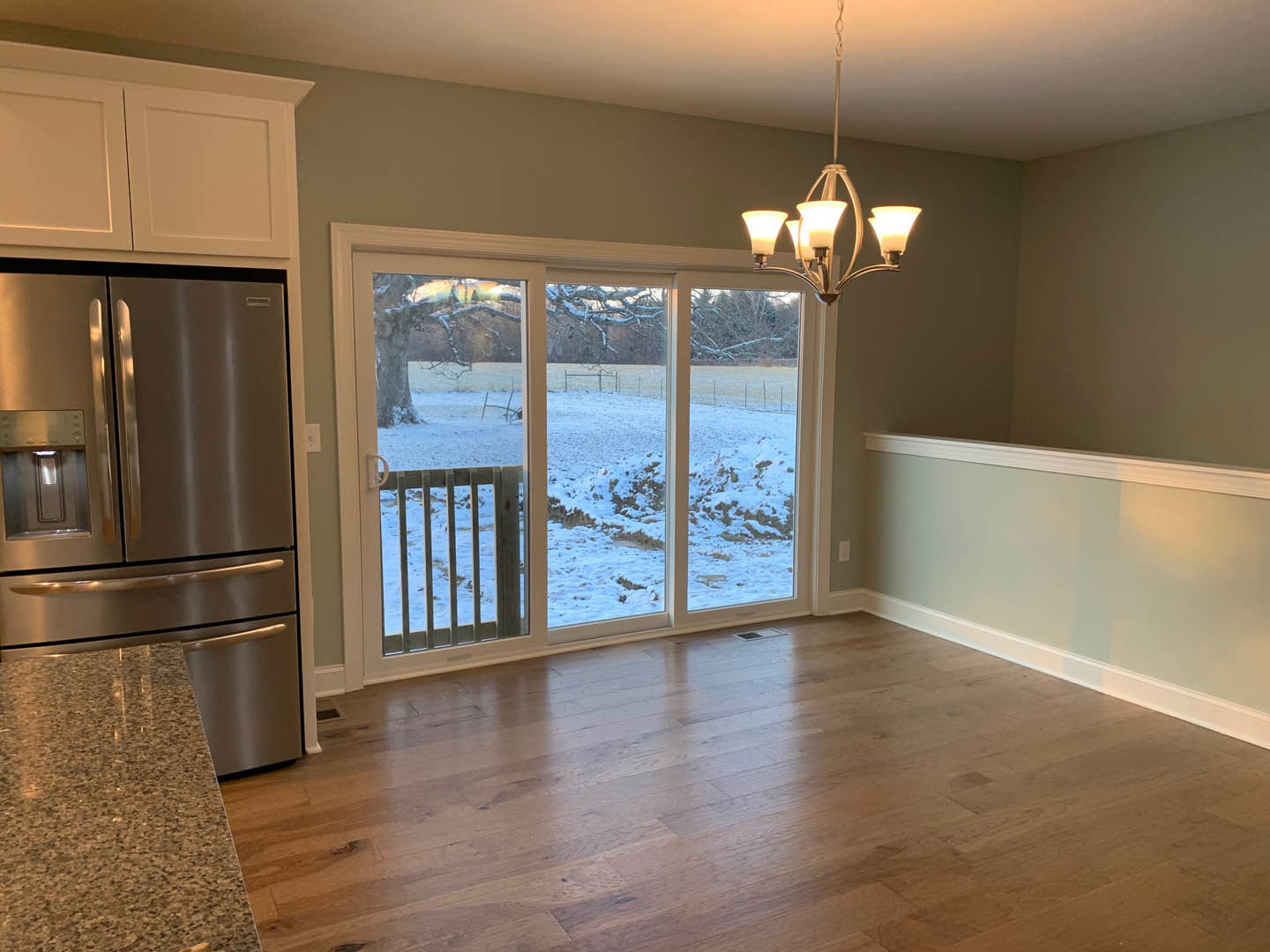 Kitchen with wood flooring, stainless steel refrigerator with ice and water dispenser, white cabinetry, sliding glass door opening to snowy yard, pendant lamp above countertop.