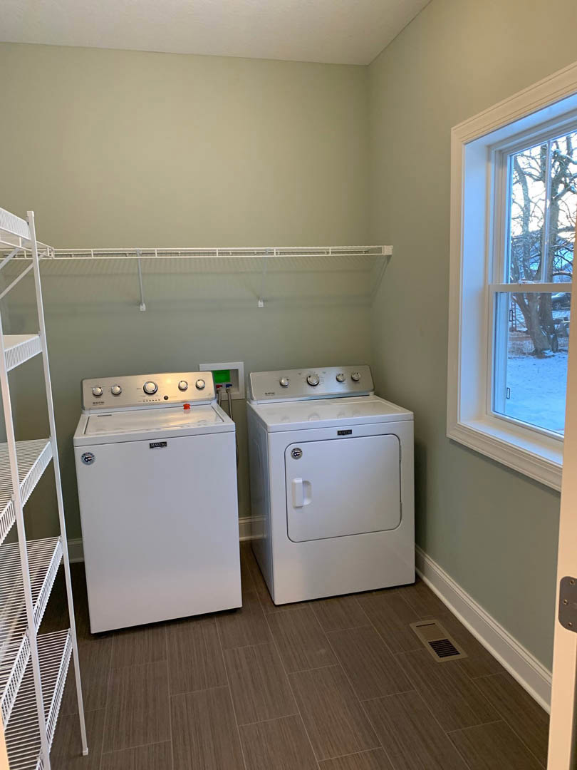 Laundry room with front-loading washer and dryer, small white refrigerator with glass top, white shelving beside washing machine, window showing tree outside, and white metal box