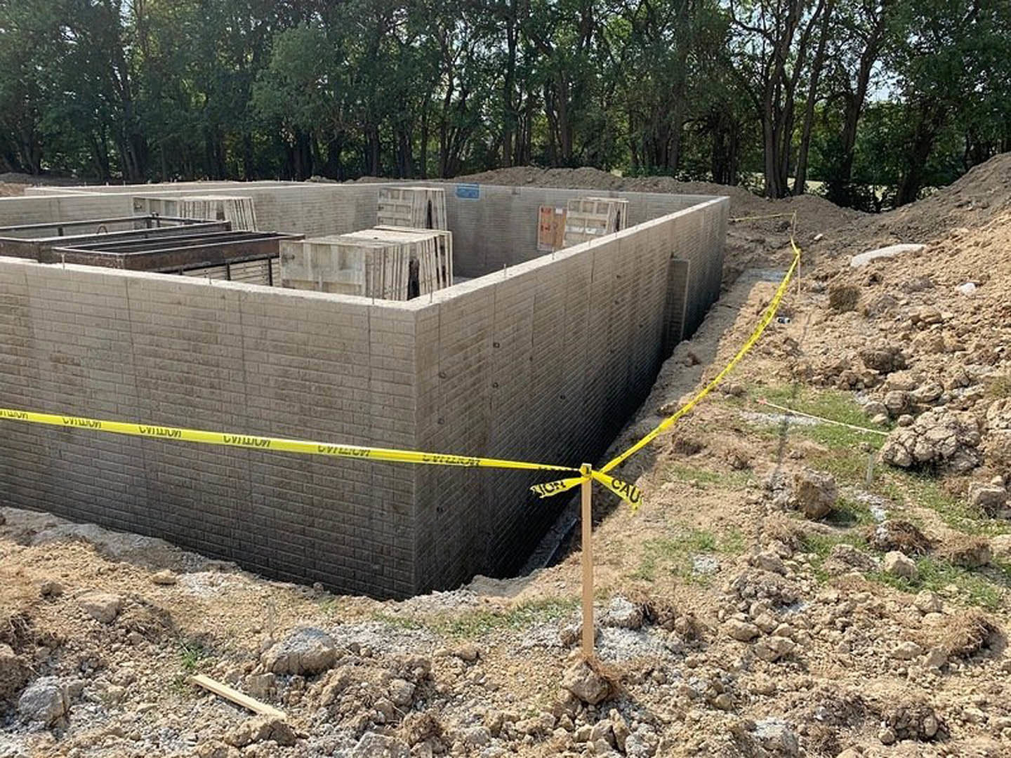 Excavated foundation surrounded by dirt, yellow caution tape, stacked concrete blocks, metal container with rebar, and trees in the background