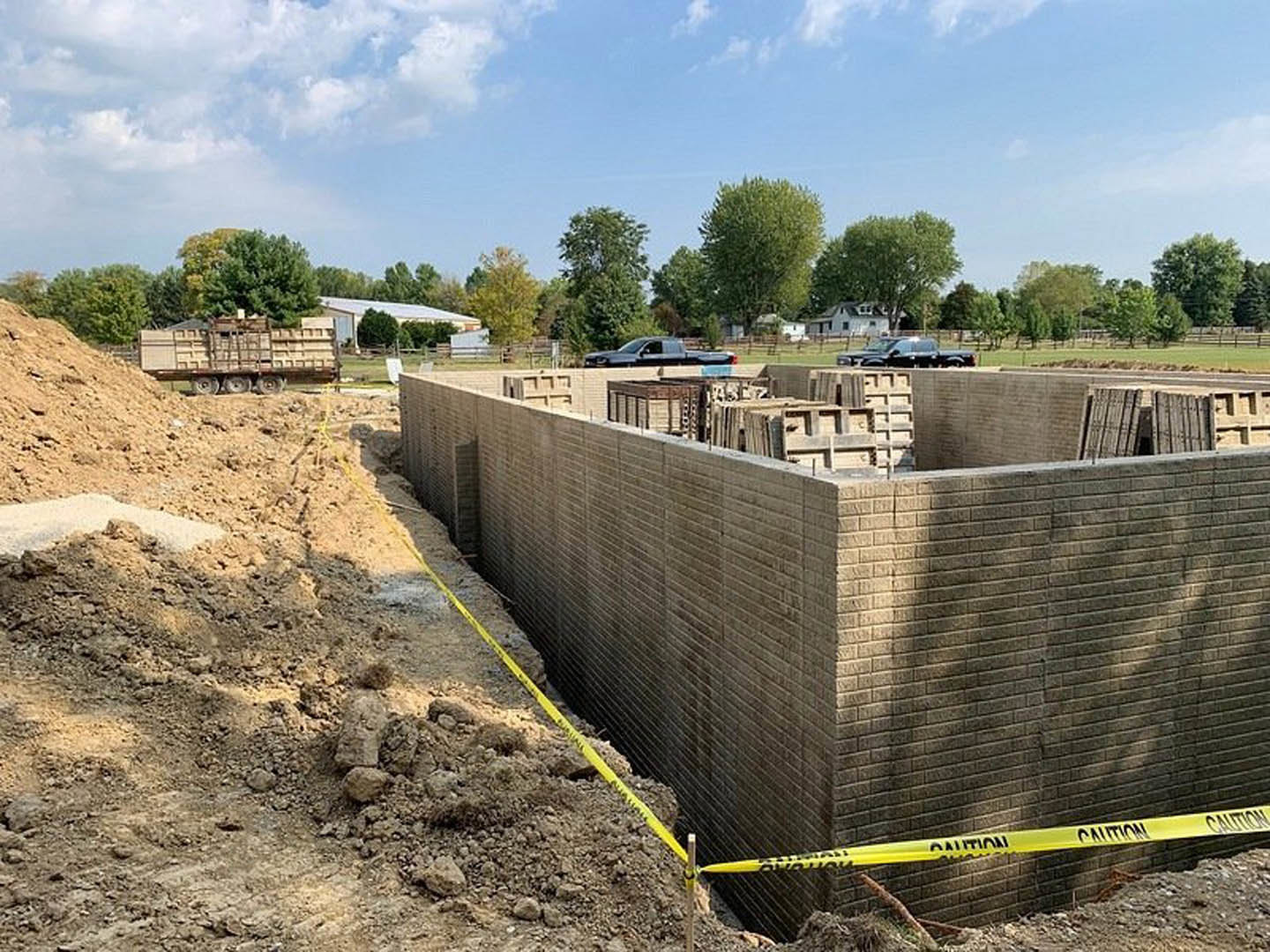 Brick wall under construction with yellow caution tape, exposed soil foundation, construction truck nearby, and trees against a blue sky with clouds in the background