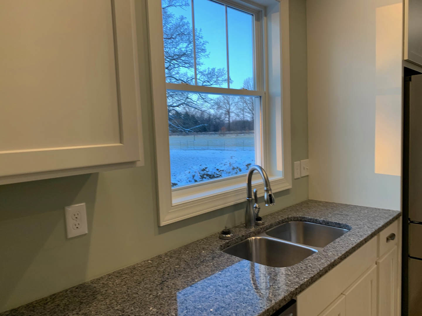 White kitchen sink with chrome faucet set in quartz countertop, outlet on backsplash, large window overlooking snowy field and trees, white cabinetry below