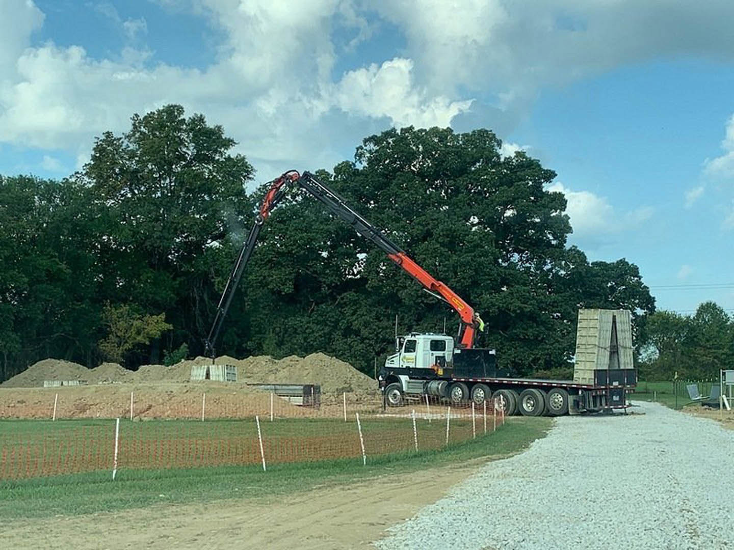 Crane lifting a truck on gravel road beside grassy land lot, blue sky with scattered clouds and trees in background