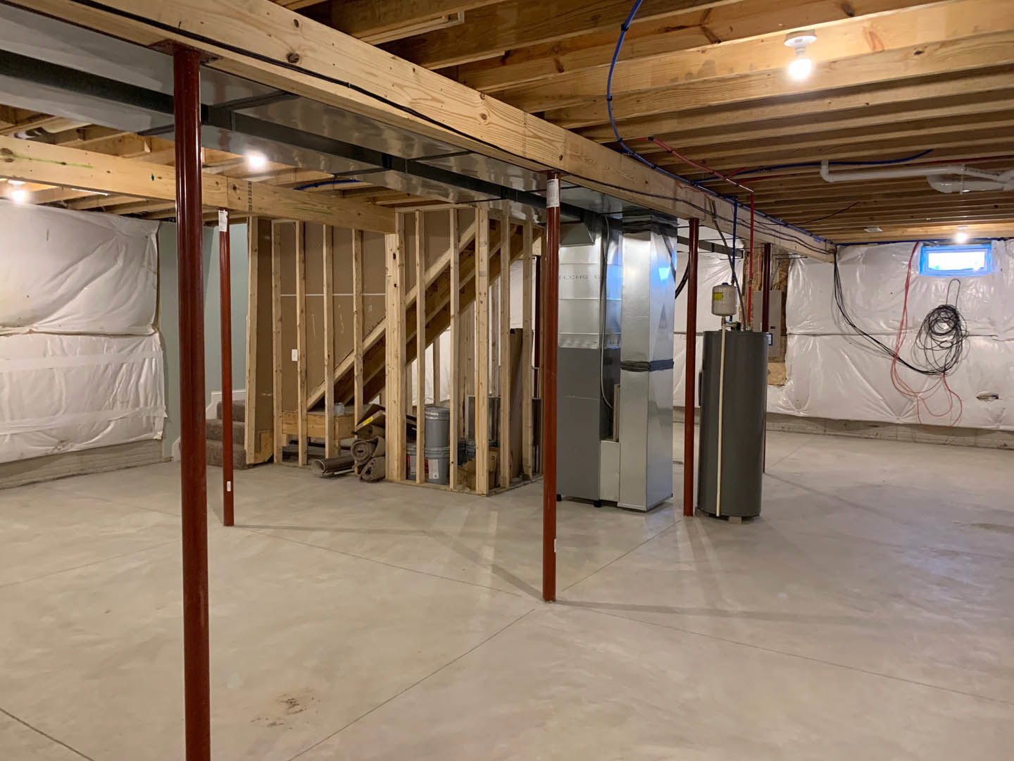 Concrete basement with exposed ceiling beams, metal staircase, steel utility box mounted on wall, and white floor with metal pole and shelving unit
