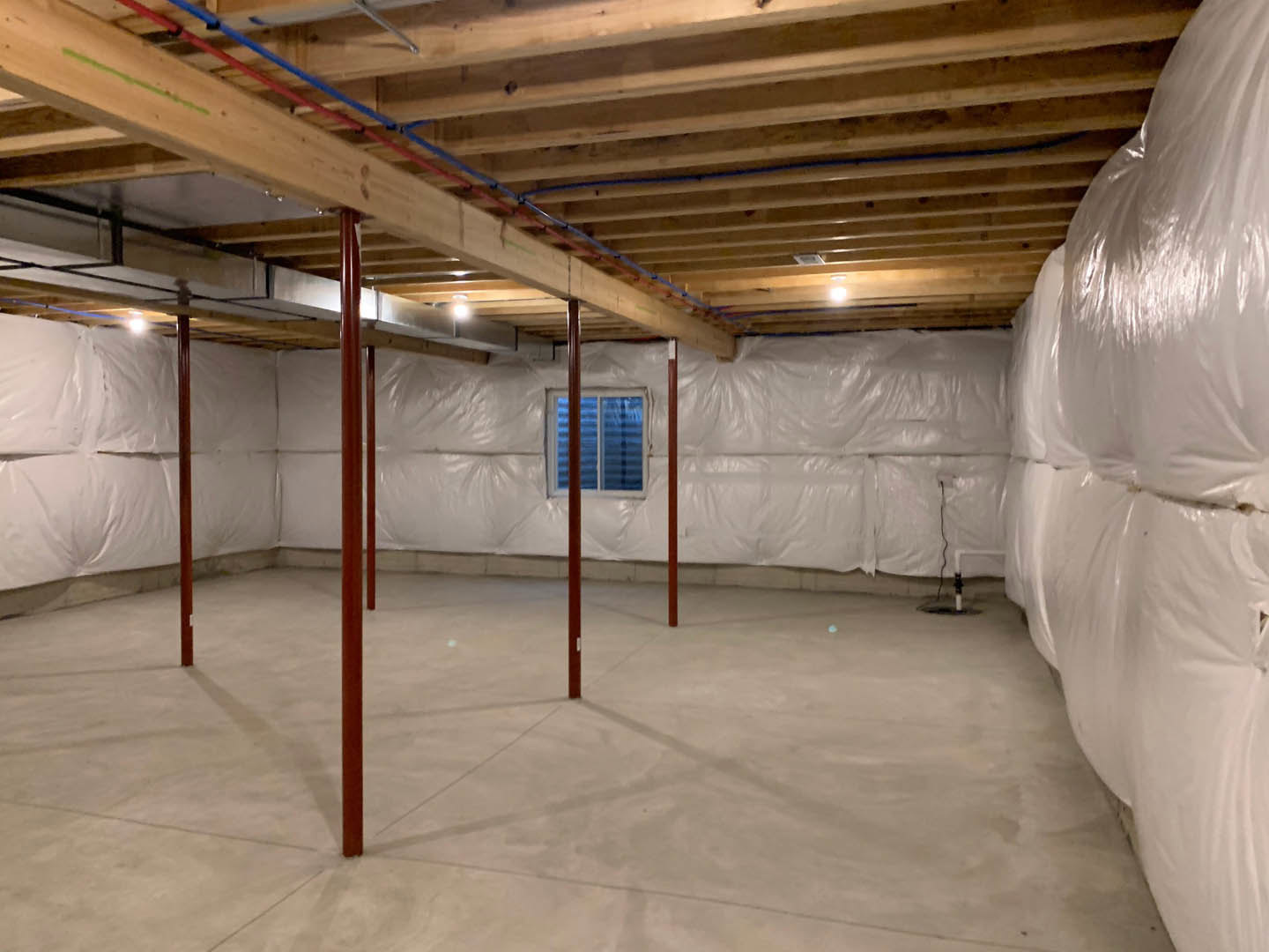 Unfinished room with exposed wooden ceiling beams, white plastic coverings on walls, concrete floor, red support poles, stack of white bags, and visible wiring