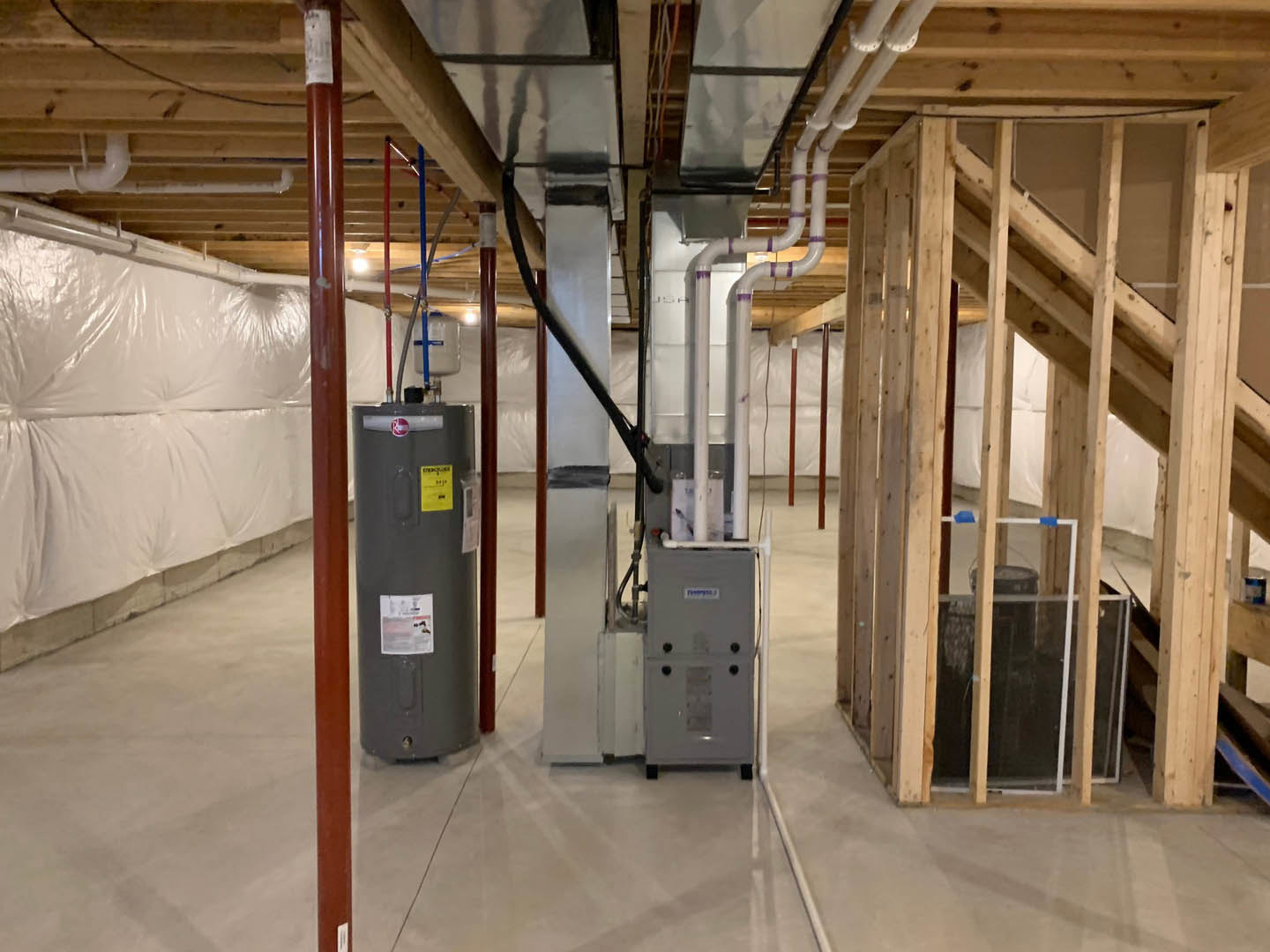 Utility room with exposed steel beams, grey walls, water heater featuring black cylinder and yellow label, visible pipes, and aluminium fixtures.