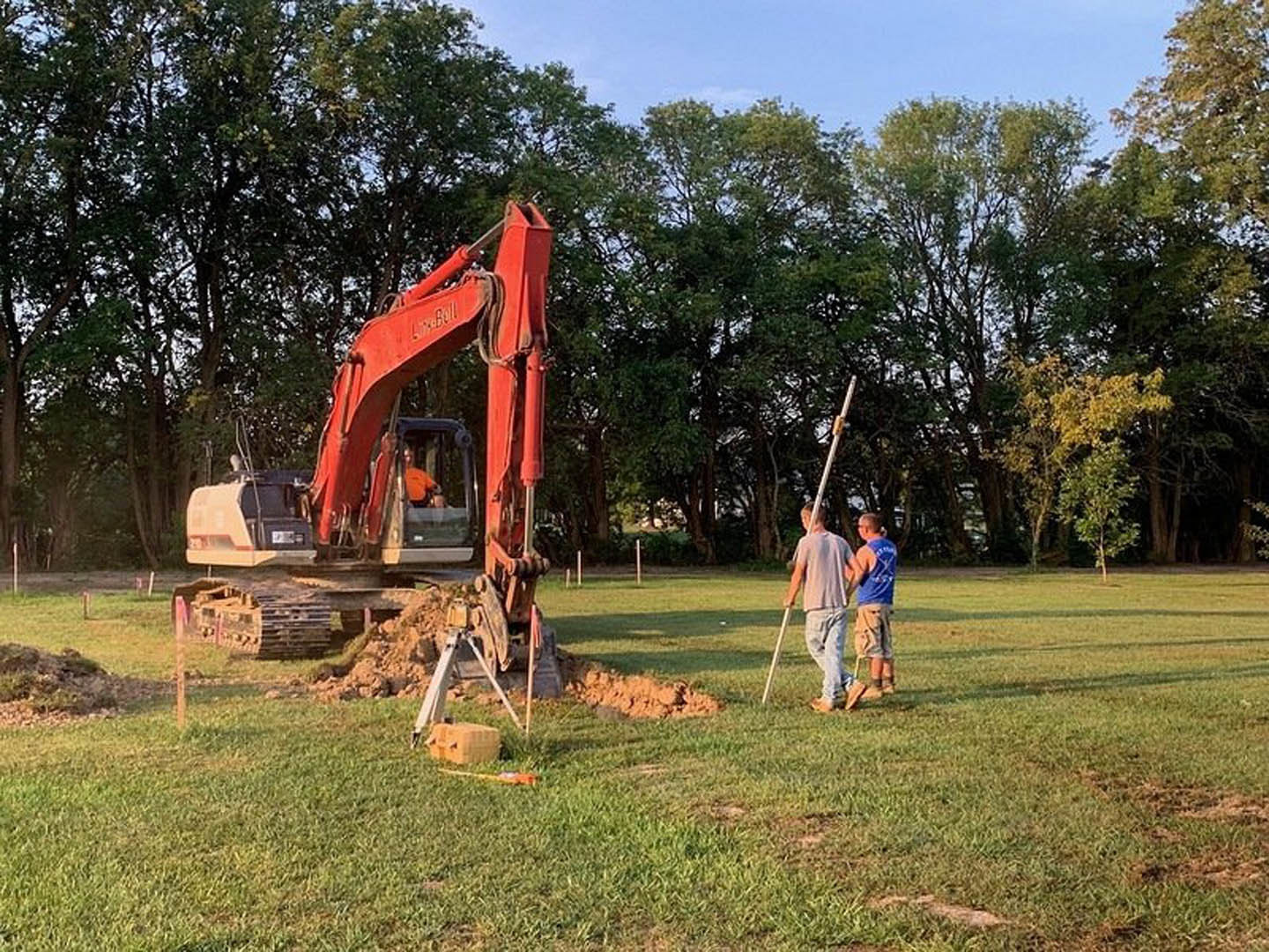 Men standing beside a red and white excavator on grassy lawn, surrounded by trees and construction materials, with blue sky overhead