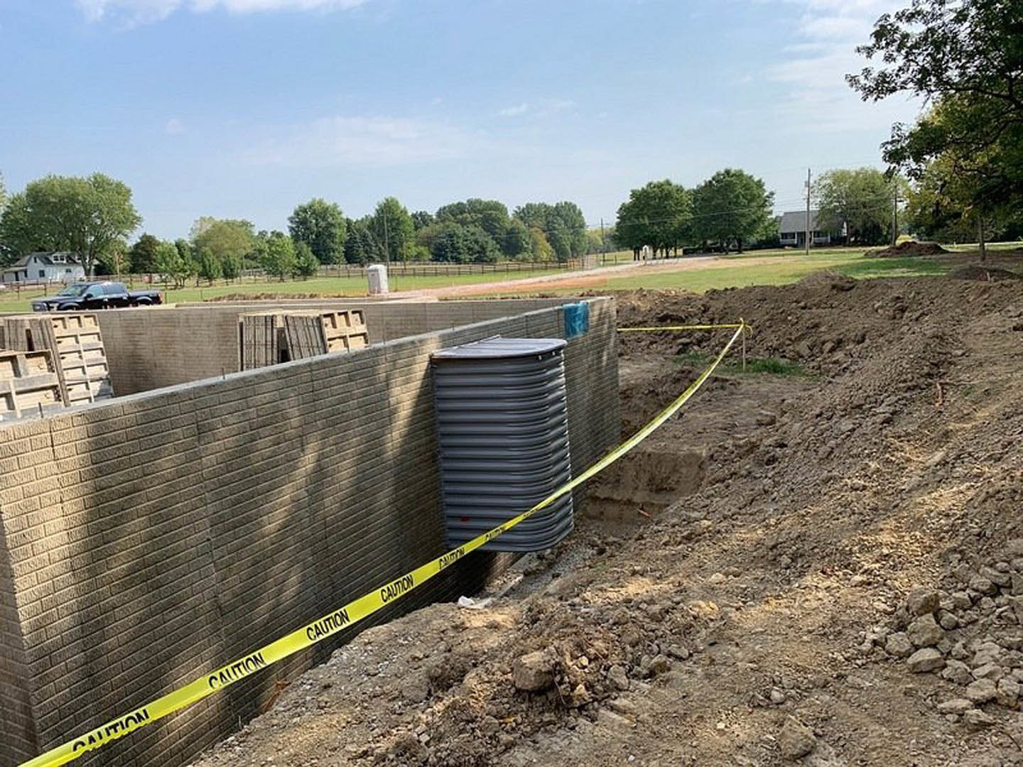 Yellow caution tape stretched across a construction site with exposed soil, a grey tank beside a brick wall, black plastic pipe, leafy tree, and scattered rocks.