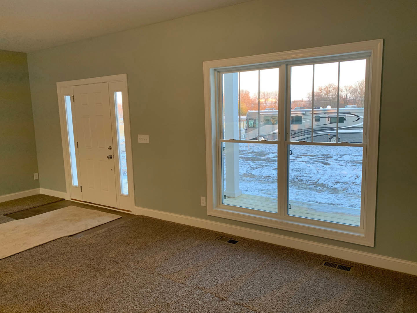 White-painted room with a single window overlooking a parking lot, white door with glass insert, neutral carpet flooring, and simple ceiling.