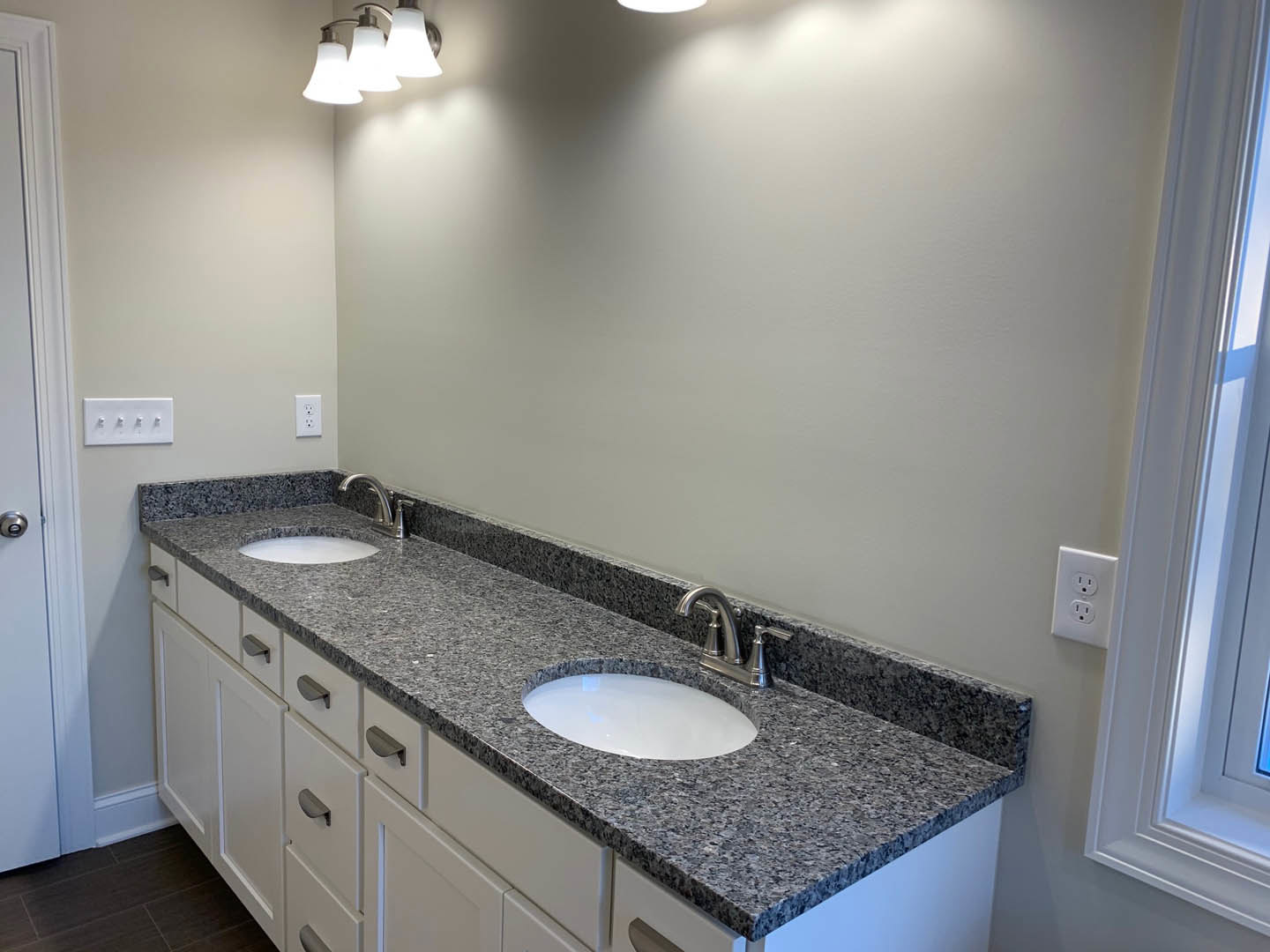 Granite countertop with undermount sink, chrome faucet, white electrical outlet, wall-mounted light fixture, large mirror, and light gray tile backsplash