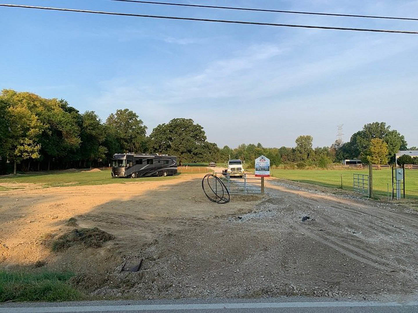 Black RV with tinted windows parked on a dirt field, surrounded by grass, trees, power lines, and a fence; sign with an image and metal ring visible in foreground.