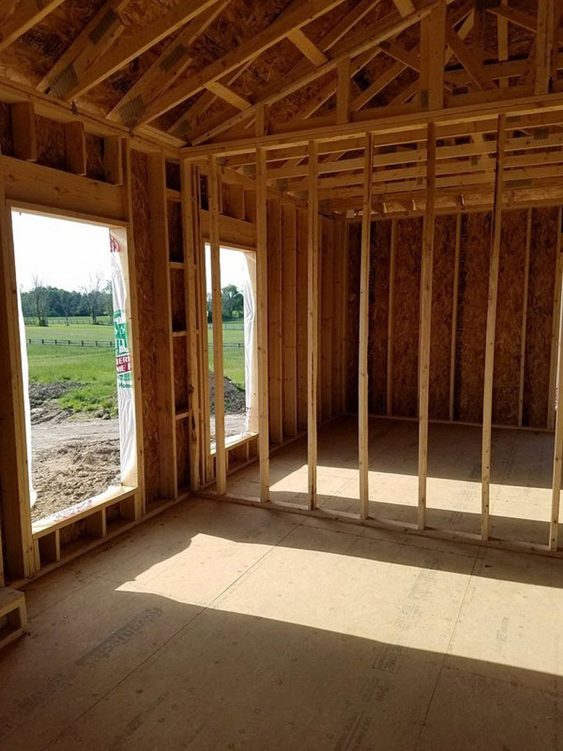 Wood-framed room under construction with exposed ceiling beams, unfinished walls, and a window overlooking a grassy field