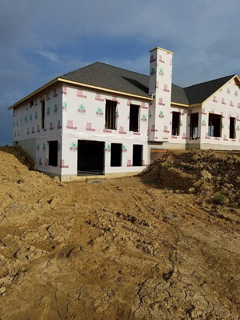White custom home under construction with red framing accents, black front door with white trim, exposed chimney, large dirt patch in foreground, blue sky and scattered clouds