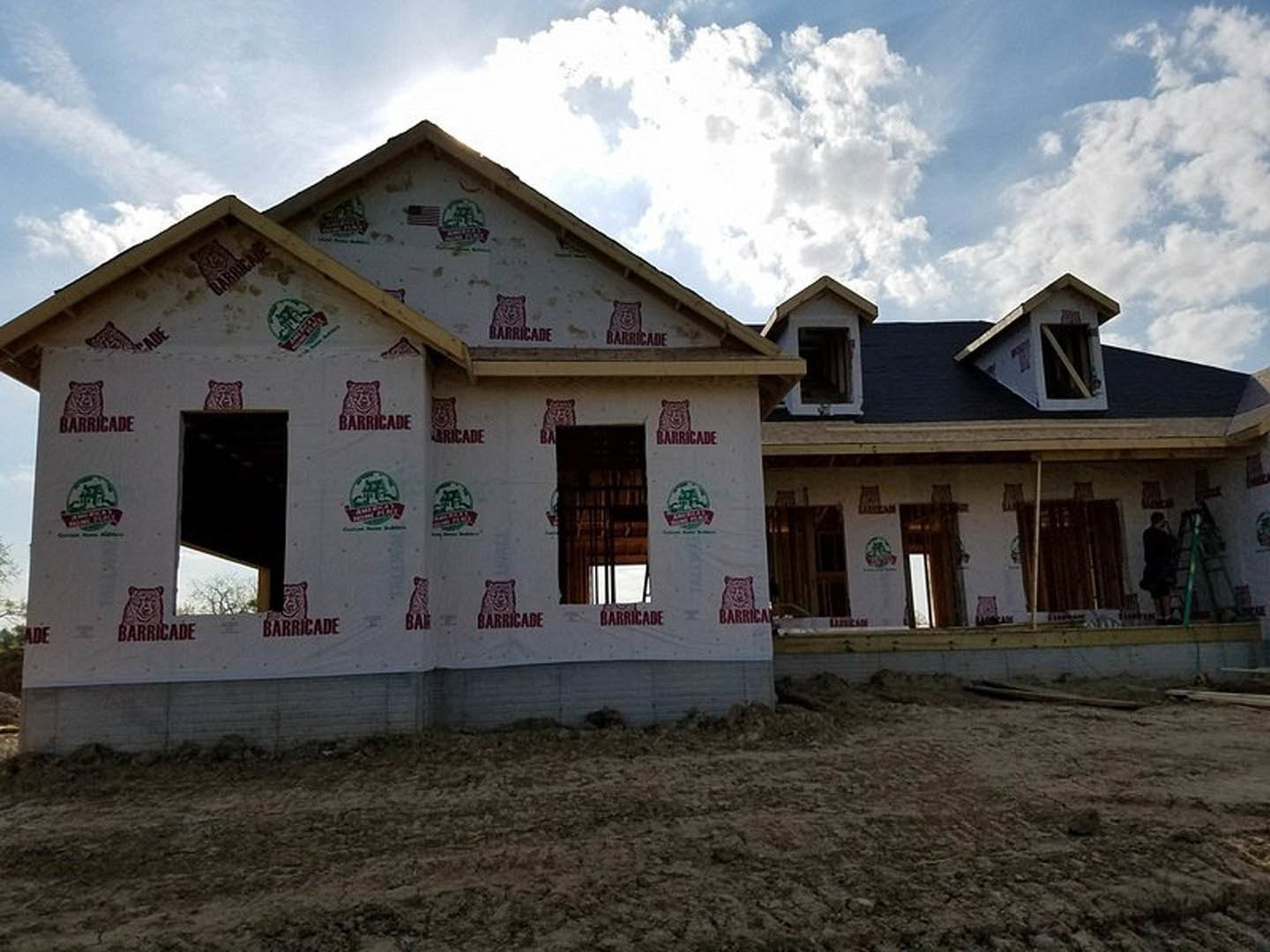 Two-story house under construction with exposed framing, several installed windows, and a patch of bare dirt in the foreground