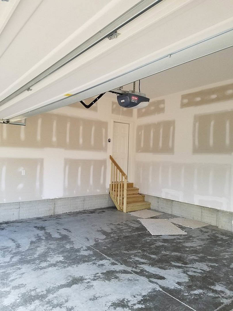 Garage interior featuring a wooden staircase with handrail, concrete floor, white ceiling with garage door opener, and white and black tile accents.