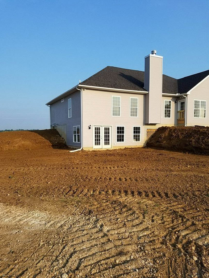 Framed house under construction with glass-paneled double doors, large dirt mound in foreground, exposed plywood walls, and open window openings